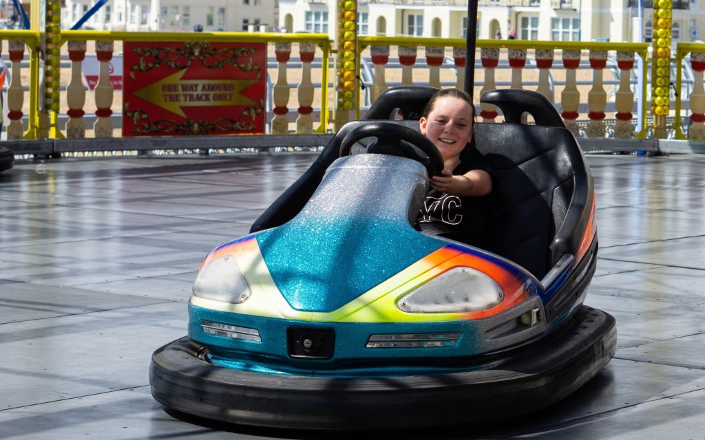 A smiling child drives a glittery blue dodgem car on an amusement park ride at Kidz Island, South Parade Pier, Southsea, surrounded by colourful decorations and safety barriers in the background.