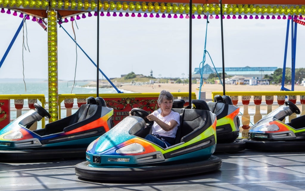 An older woman drives a colourful dodgem at Kidz Island, an outdoor amusement park on South Parade Pier in Southsea, with the sandy beach and sea visible in the background.