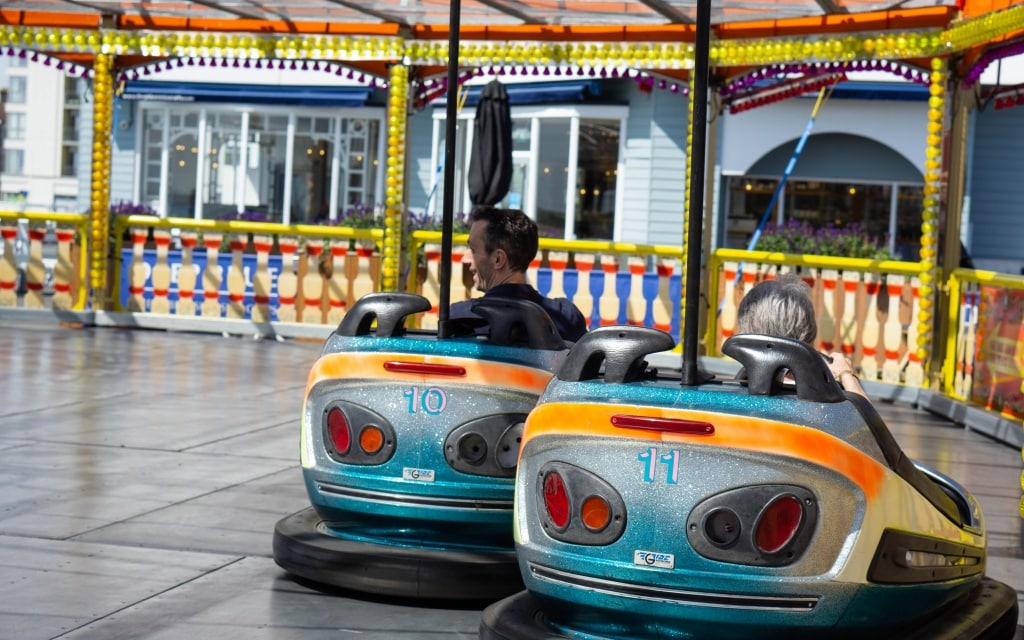 Two people ride dodgems numbered 10 and 11 at Kidz Island, a colourful attraction at South Parade Pier in Southsea. The area features bright lights and railings, with the cars stationary on the ride platform.