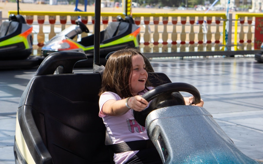 A young girl with long brown hair smiles and steers a dodgem at Kidz Island near South Parade Pier on a sunny day. Other colourful dodgems and a fence are visible in the background at this Southsea amusement park ride.
