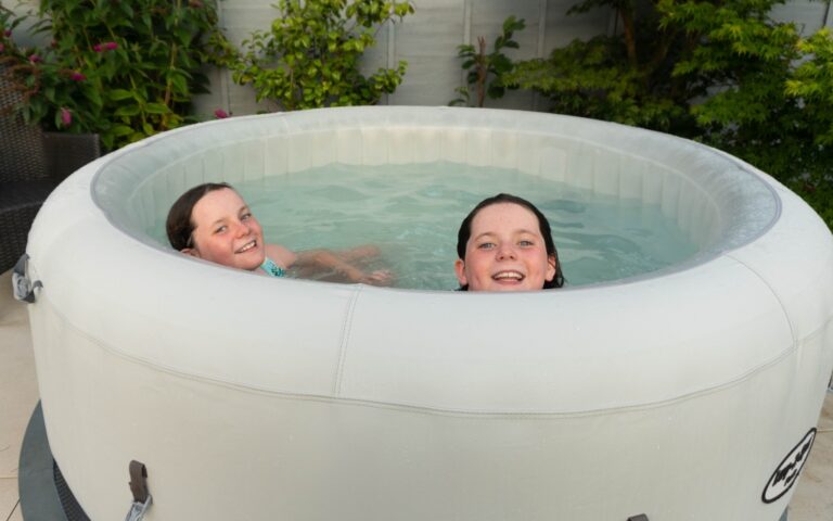 Two children smile while relaxing in a Lay-Z-Spa Paris hot tub outdoors, surrounded by green plants and a fence. Only their heads and shoulders are visible above the water, making it an inviting scene for any hot tub review.