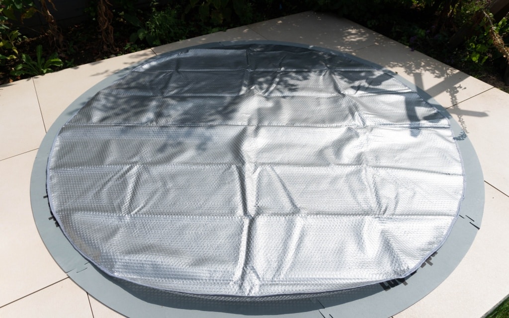 A round, silver bubble wrap pool cover is spread out on a tiled patio beside a Lay-Z-Spa Paris hot tub, partially shaded by surrounding plants.