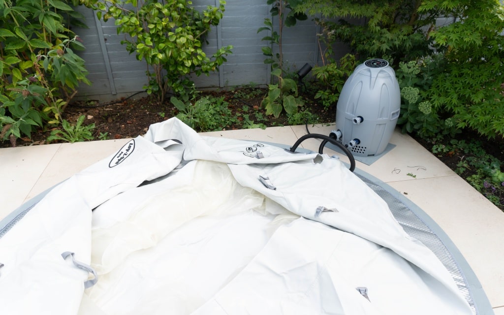 A deflated Lay-Z-Spa Paris hot tub is laid out on a patio in a garden, with a grey pump or heater unit placed nearby among green plants and shrubs, ready for the next relaxing soak or review.