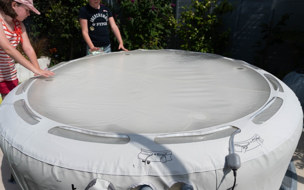 Two children stand near a Lay-Z-Spa inflatable hot tub covered with a fitted cover. One wears a red striped top and hat, the other a dark t-shirt. The hot tub is outdoors, surrounded by plants and a fence.
