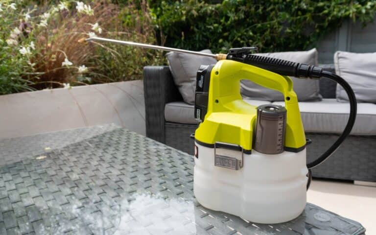 A yellow and white Ryobi cordless weed sprayer sits on a glass outdoor table, with garden furniture and green plants visible in the background.