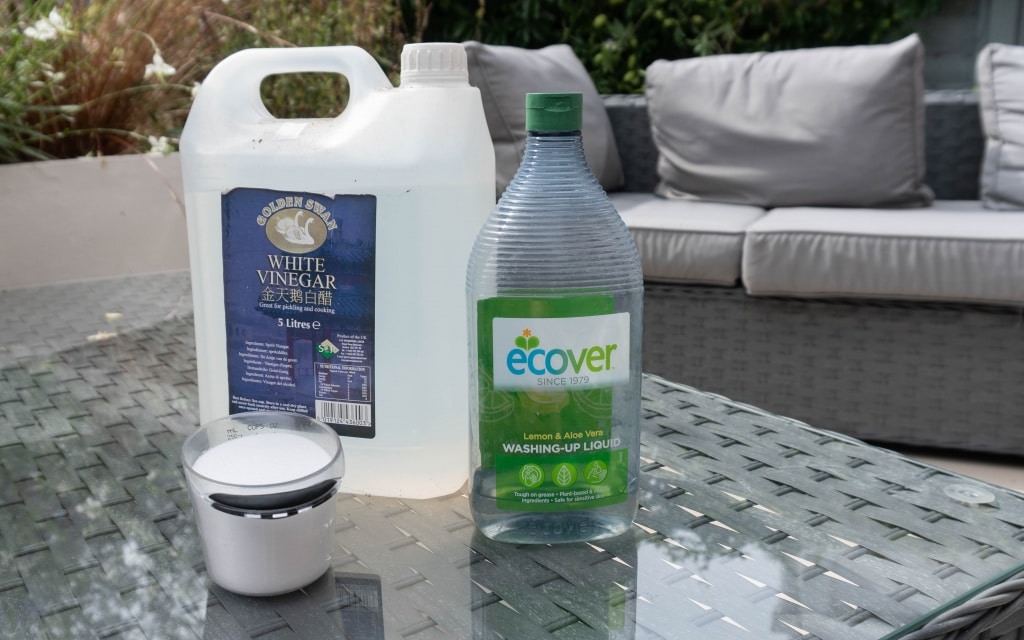 A bottle of white vinegar, Ecover washing-up liquid, and a small white container sit on a glass outdoor table—perfect for mixing solutions for your Ryobi Weed Sprayer. A grey patio sofa and lush plants complete the scene.