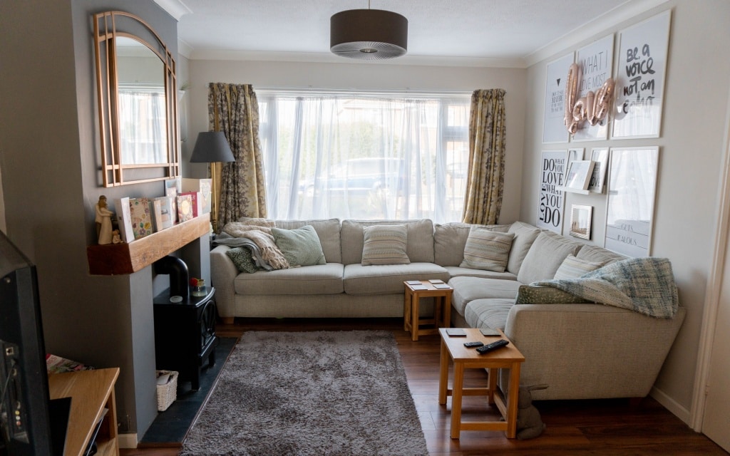 A cosy living room in this house tour features a large L-shaped beige sofa, two wooden side tables, a fireplace with a mantelpiece, wall art, a grey rug, and large windows with curtains letting in natural light. This inviting space is part of an extension to the existing house.