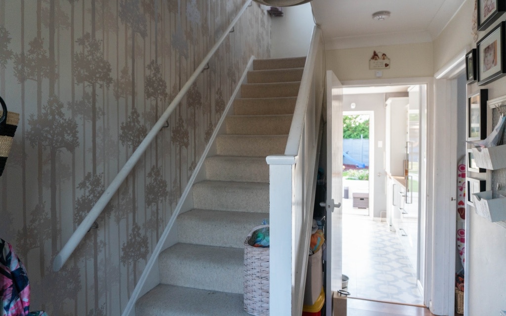 A hallway with a carpeted stairway on the left, floral-patterned wallpaper, and a basket at the foot of the stairs. Sunlight from an extension streams in through an open doorway at the end—an inviting scene from our existing house tour.