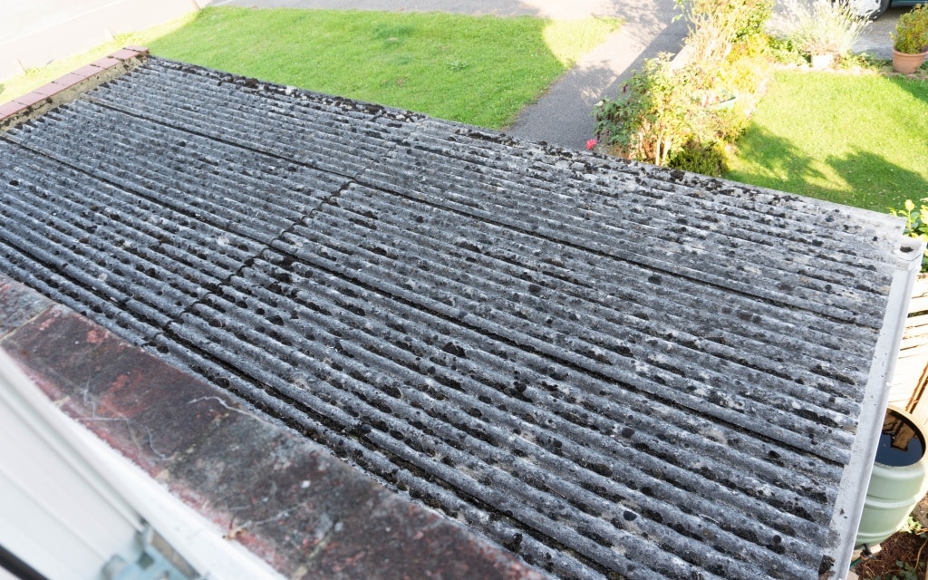 A corrugated asbestos roof with visible dirt, moss, and weathering, viewed from above during an Existing House Tour, with a green lawn, plants, and part of a drive in the background.