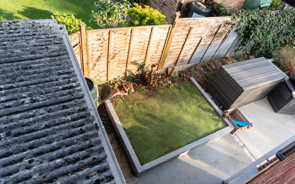 View from above of a small back garden behind the existing house, featuring a square lawn bordered by concrete, a wooden fence, patio furniture, and a paved path. Some plants and garden tools are visible near the fence.