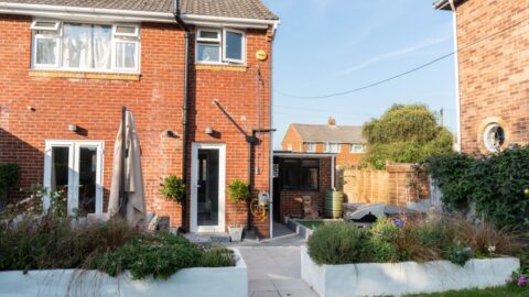 A red-brick, two-storey house with white-framed windows and doors opens onto a tidy back garden patio, where a house extension blends seamlessly with raised flower beds, plants, and garden furniture on a sunny day.