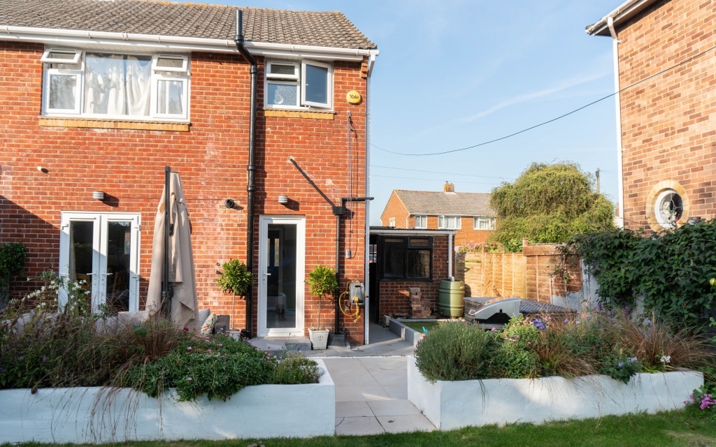 A red-brick, two-storey house with white-framed windows and doors opens onto a tidy back garden patio, where a house extension blends seamlessly with raised flower beds, plants, and garden furniture on a sunny day.