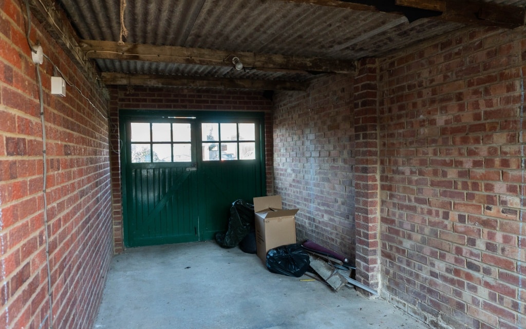 A mostly empty garage with red brick walls, a concrete floor, and a green double door with windows. Part of an extension to the existing house tour, it features two black bin bags, a cardboard box, and some miscellaneous items in one corner.