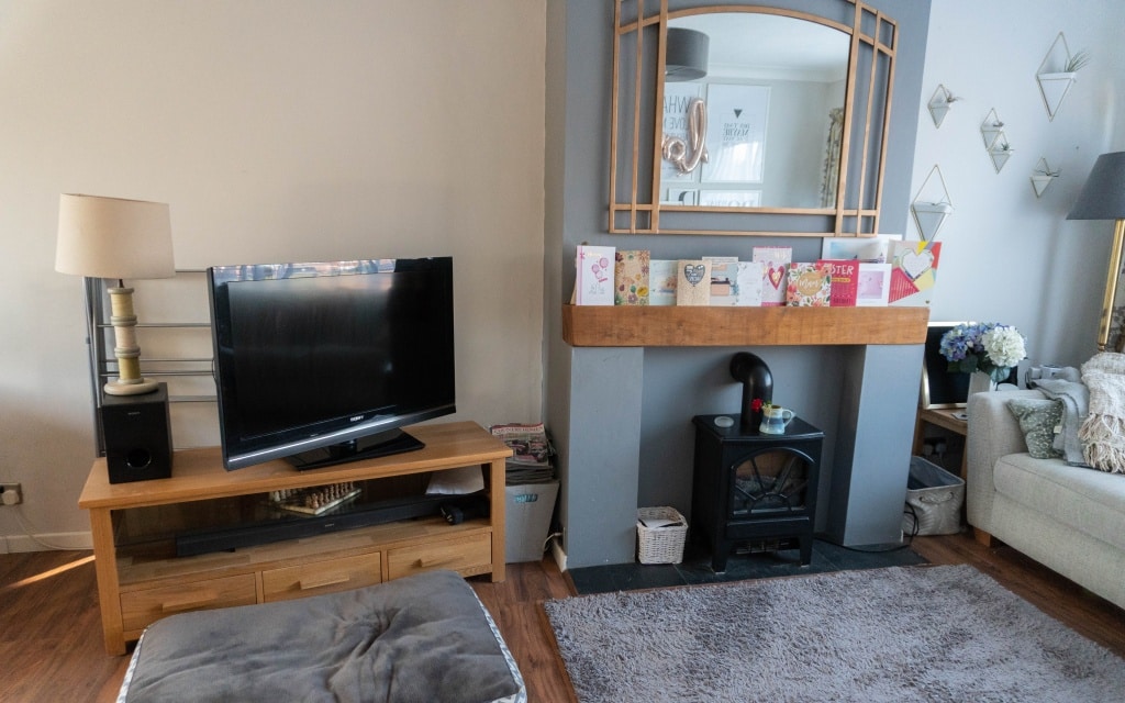 A cosy living room with a TV on a wooden stand, a fireplace decorated with cards, and a large mirror above the mantelpiece offers inviting charm in this existing house tour. A sofa, cushions, soft ottoman, light decor, and wooden flooring complete the look.