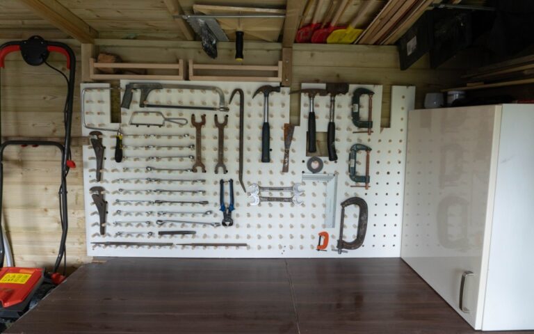 A white pegboard mounted on a wooden wall showcases neat shed organisation, holding saws, clamps, screwdrivers, and spanners. A white cupboard stands on the right, while a red lawnmower is partially visible on the left.