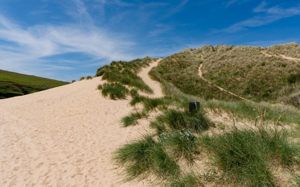 A sandy dune with patches of green grass under a blue sky with wispy clouds at Holywell Bay Beach. Walking trails wind up the dune, and a small black sign is visible among the grass near Newquay.