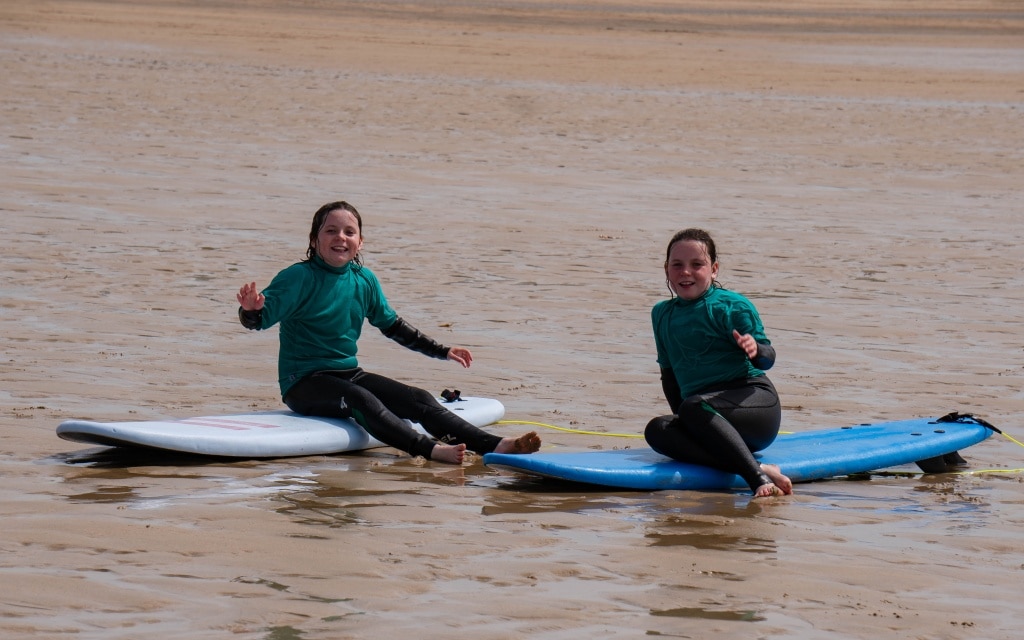 Two children in wetsuits sit on surfboards on wet sand at Holywell Bay Beach, smiling and waving at the camera. The tide is low, leaving a wide expanse of sand and shallow water around them near Newquay.
