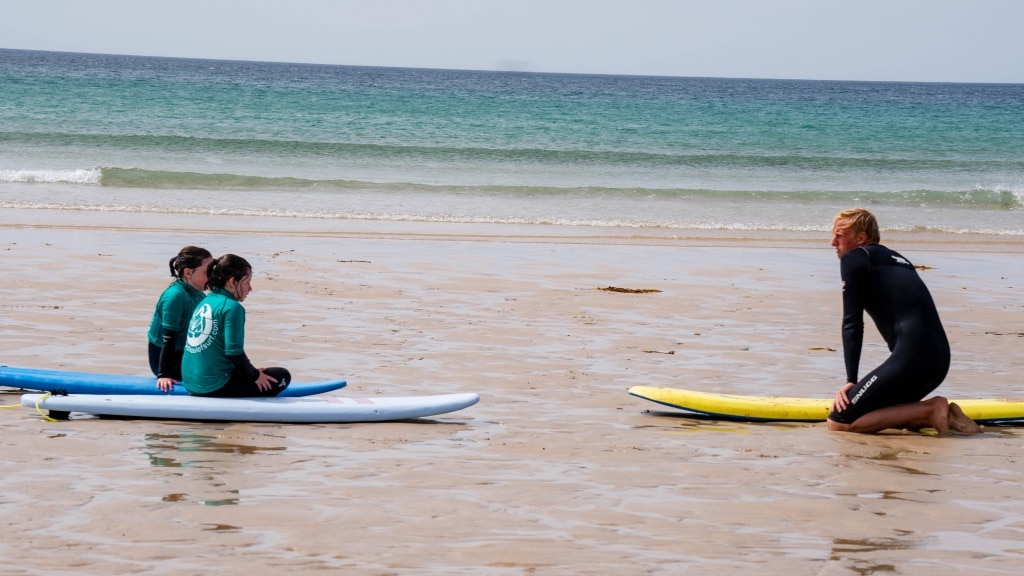 Two people sit on surfboards on the wet sand at Holywell Bay Beach while a surf instructor in a wetsuit kneels on a yellow surfboard, giving instructions. The sea and sky are visible in the background.