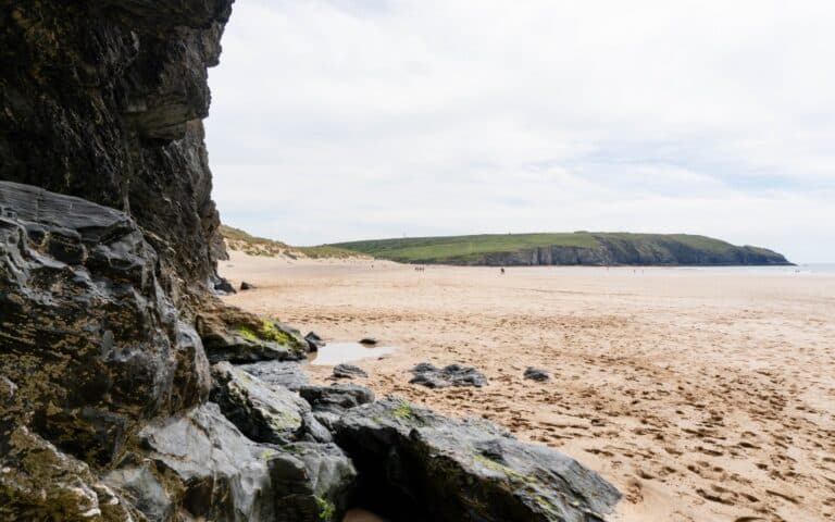 A sandy beach with footprints, bordered by rocky cliffs on the left, captures the charm of Holywell Bay near Newquay. In the distance, green hills rise under a cloudy sky, with few people visible far away on the shore.