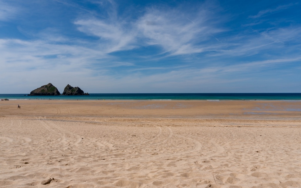 A wide sandy beach at Holywell Bay stretches towards a calm blue sea under a partly cloudy sky, with two rocky islets visible in the distance. Subtle tyre tracks and footprints mark the sand near this scenic Newquay coastline.