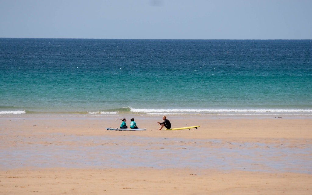 Three people sit on surfboards on the sandy beach at Holywell Bay near the shoreline, with calm blue and green sea waves in the background under a clear sky.