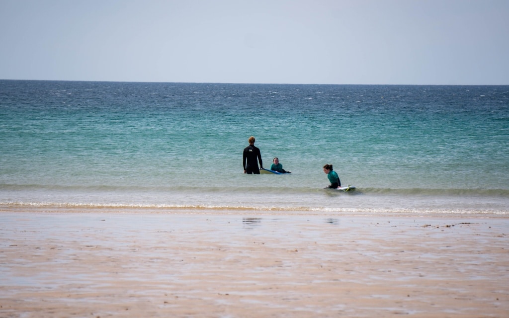 Three people in wetsuits enjoy the calm sea at Holywell Bay Beach near Newquay; two stand in the shallow water while one lies on a surfboard close to the shore under a clear sky.