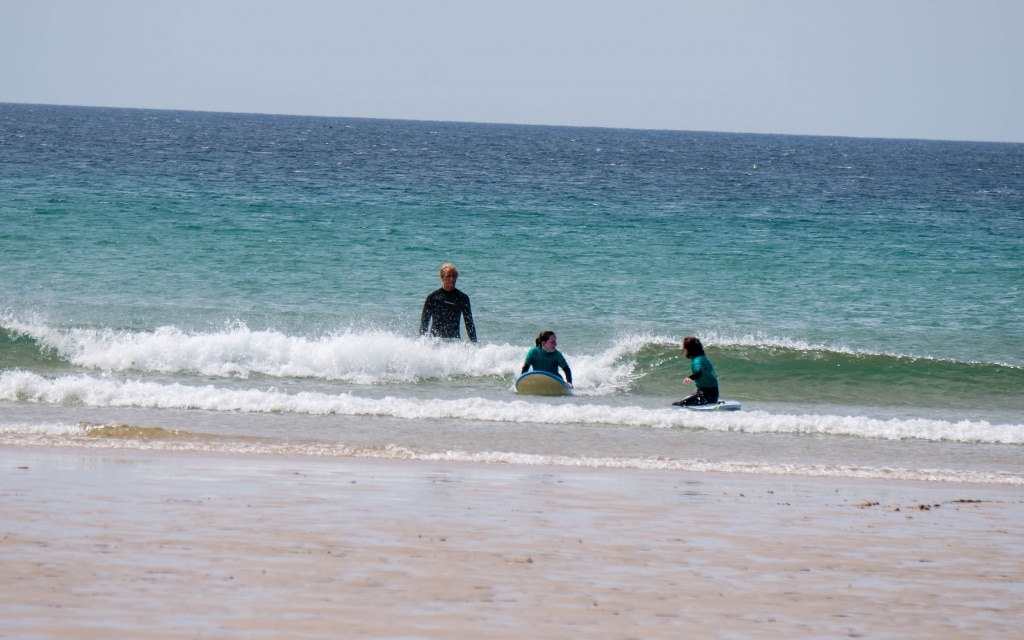Three people in wetsuits are in shallow sea waves near the shore at Holywell Bay Beach, with two sitting on surfboards and one standing in the water, sandy beach in the foreground, and calm sea stretching towards Newquay in the background.