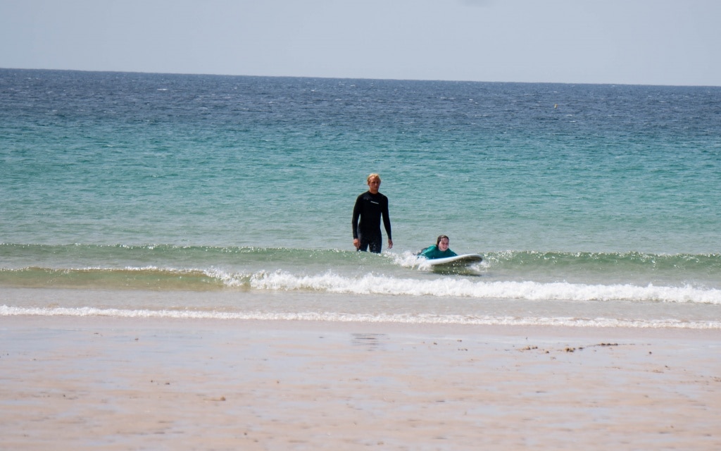 A person in a wetsuit stands in shallow sea water at Holywell Bay near a child lying on a surfboard, riding a small wave towards the sandy shore.