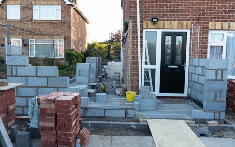 A house extension under construction features partially built brick walls, stacks of bricks, a yellow bucket, and building materials on the floors near a black front door. Neighbouring houses and a garden appear in the background.