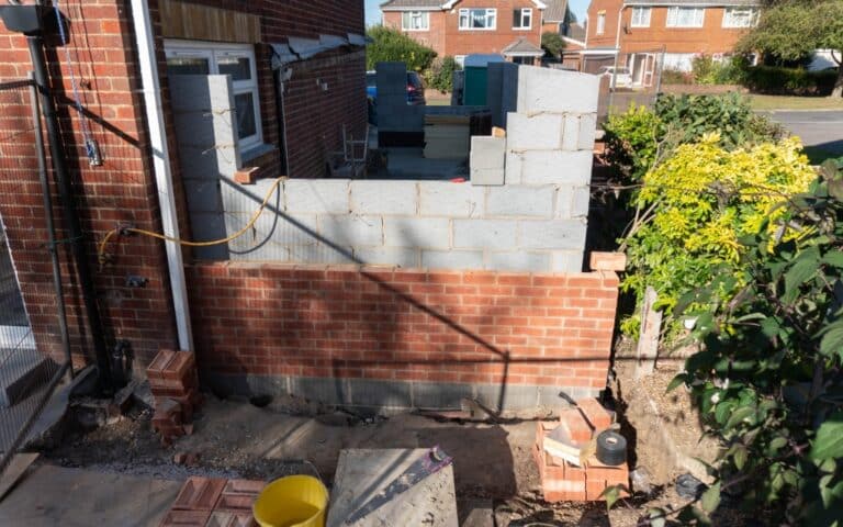 A partially built house extension features walls of grey concrete blocks and red bricks. Building materials and tools are scattered nearby, with plants visible to the right. The floors are unfinished, reflecting the ongoing work.