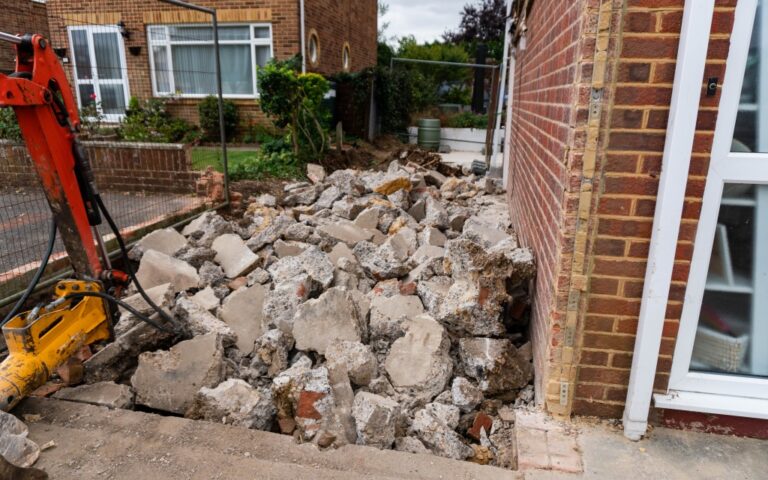 A pile of broken concrete slabs lies beside a brick house, with construction equipment and fencing nearby, marking Week 1 of a house extension project in this residential area.