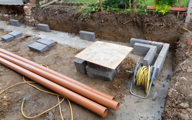 Several concrete blocks, orange pipes, and a reel of yellow hose are arranged on a soil foundation at a building site, suggesting the early stages of a house extension, with greenery and a garden area visible in the background.