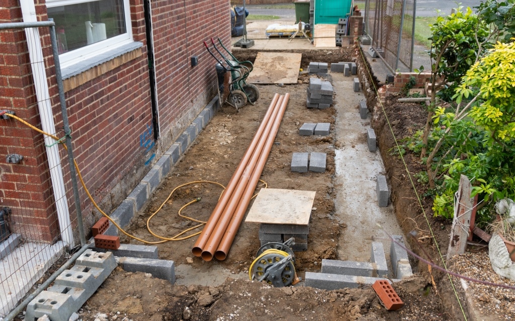 A back garden construction site beside a brick house shows week 2 of a house extension. Pipes, breeze blocks, bricks, and tools are scattered on soil with some areas dug for foundations; garden plants border the work area.