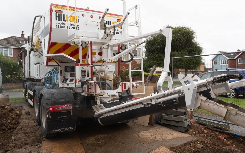 A concrete mixer lorry with an extended conveyor is pouring concrete for house extension foundations at a residential building site. Houses and trees are visible in the background, with building materials scattered nearby.