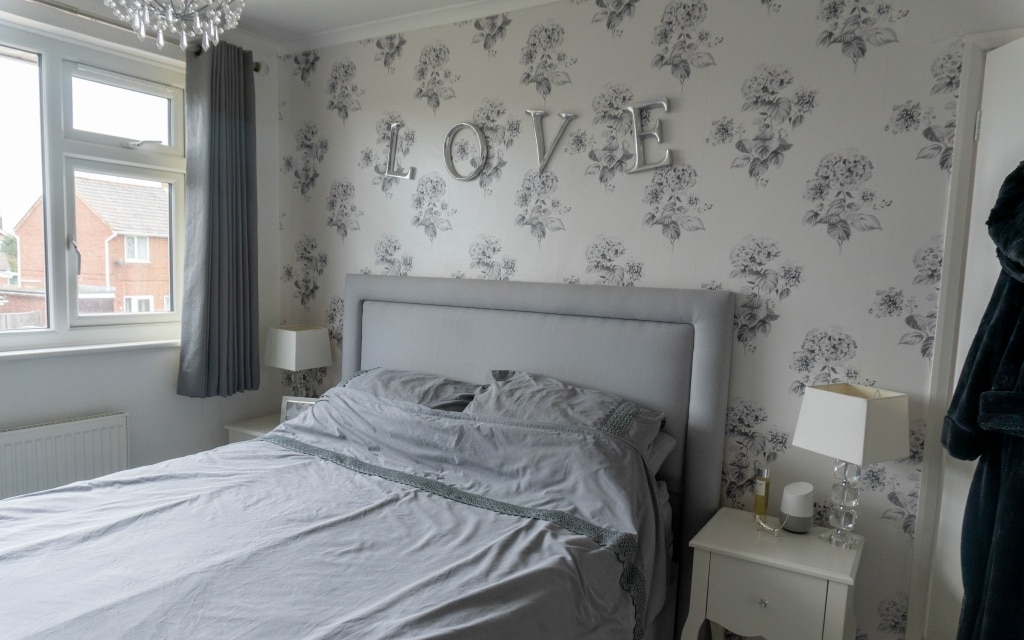 A neatly made bed with grey bedding is centred in this elegant bedroom, set against floral wallpaper. Silver LOVE letters hang above the tufted headboard, whilst Couture Living made-to-order curtains frame two white bedside tables on each side.