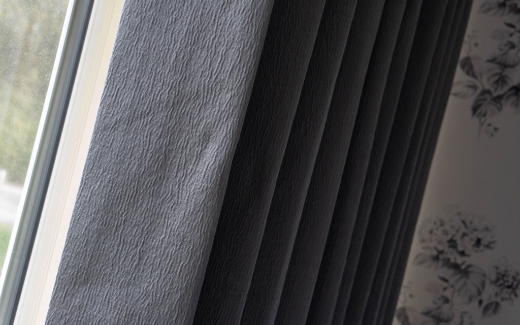Close-up view of dark grey, textured couture living bedroom curtains partially covering a window, with light filtering through on the left. The right side features a wall with floral-patterned wallpaper.