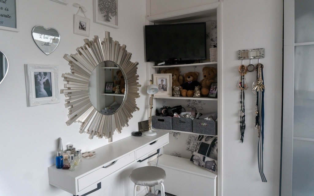 A white dressing table area with a round sunburst mirror, small stool, and shelves holding soft toys, jewellery, and framed photos offers clever bedroom storage solutions. Necklaces hang on hooks to save space, while bottles sit neatly on the dressing table.