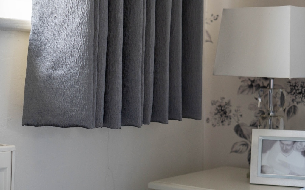 A close-up of a bedroom corner features Couture Living made-to-order curtains, a table lamp with a clear base, and a framed black-and-white photo on a white bedside table against floral-patterned wallpaper.