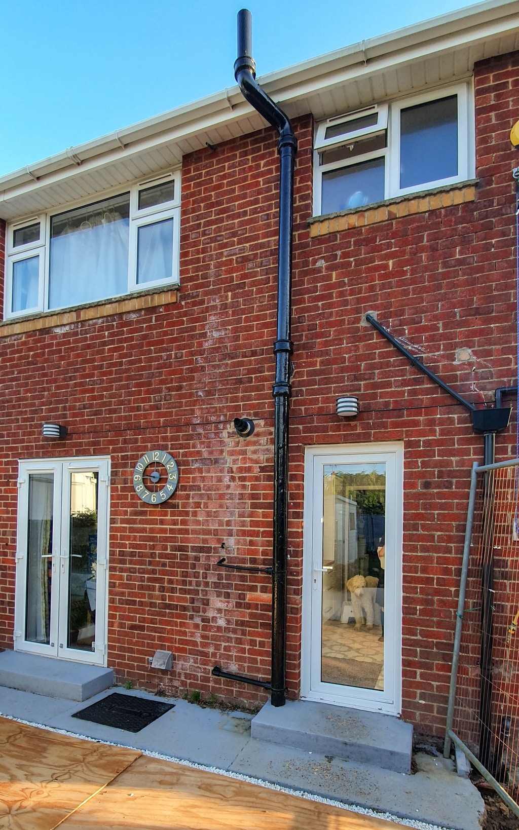 A red brick house with two white doors and several windows. There is new black pipework attached to the bricks, and a round clock next to one of the doors. A dog is visible inside behind one of the glass doors.