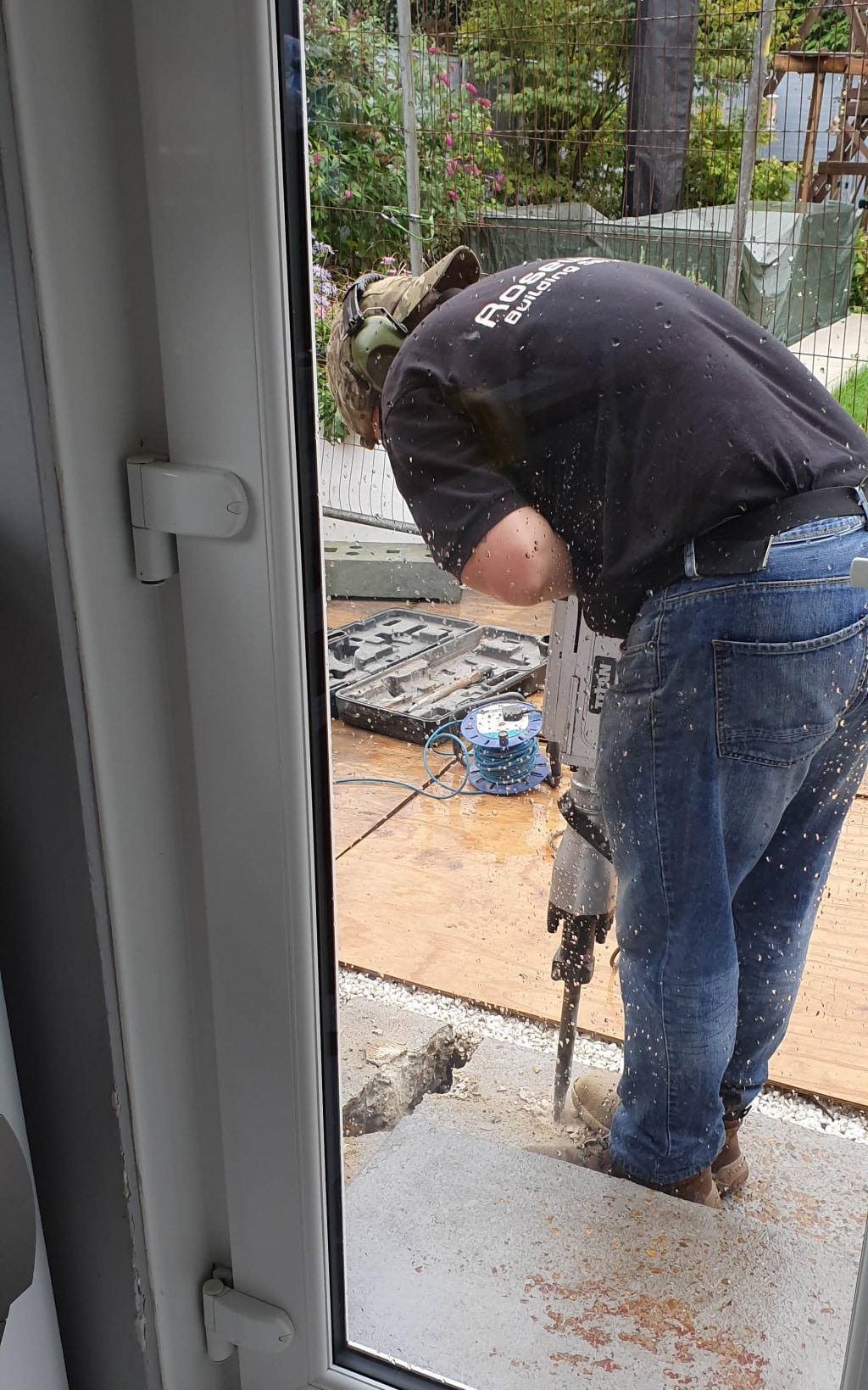 A person in jeans and a black shirt uses a power drill on a concrete surface outside, viewed through a rain-speckled glass door. Tools, cables, and bricks hint at a possible house extension project nearby.