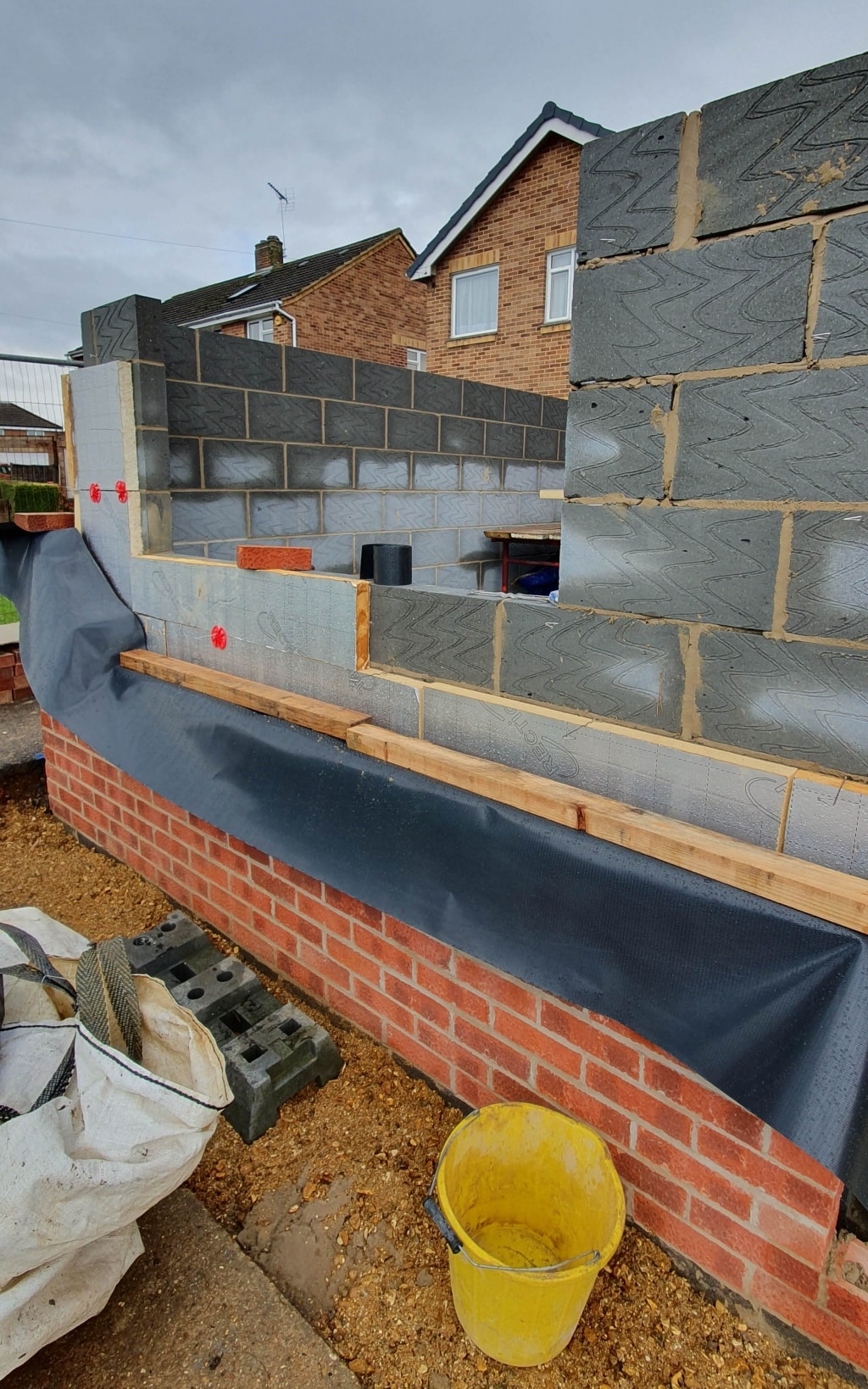 A partially constructed brick and block wall for a house extension on a building site, with insulation and a black damp-proof membrane visible. A yellow bucket and stacks of bricks are in the foreground, with a house in the background.