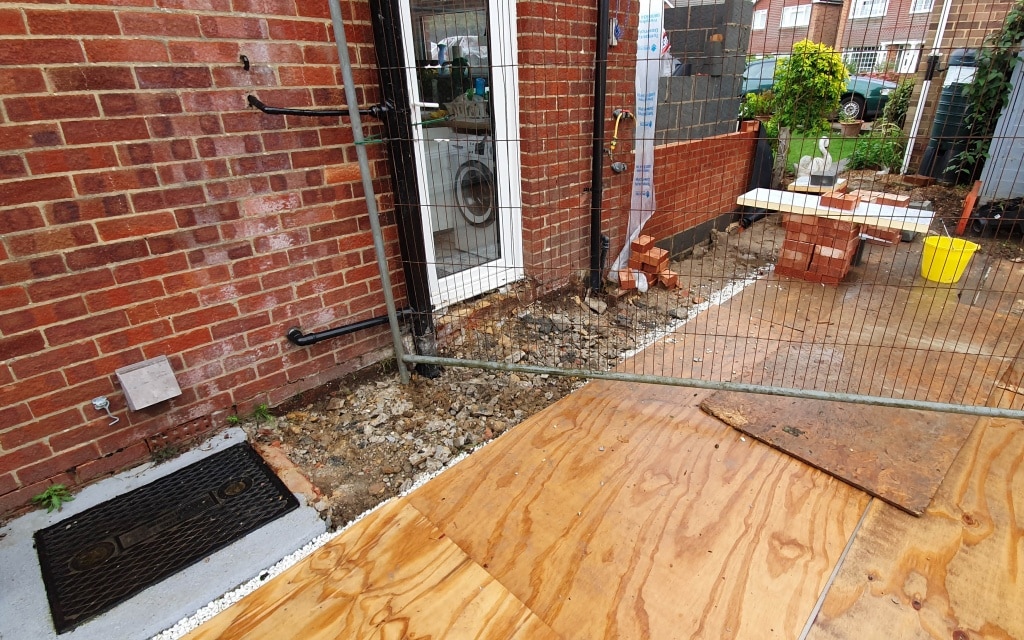 A construction site outside a brick building shows exposed ground, plywood sheets, stacked bricks for a house extension, a metal fence, and building materials near a glass door.