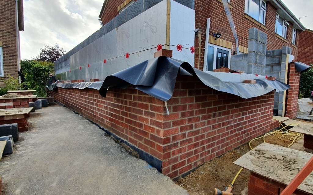A partially constructed house extension features brick walls, insulation panels, and plastic sheeting. Building materials and sand heaps are visible next to the property, along with unfinished surfaces.