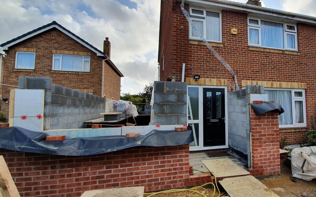 A partially constructed house extension features exposed grey blocks and visible building materials. The main house's red bricks and white-framed windows contrast with a wooden plank and yellow cables in the foreground.
