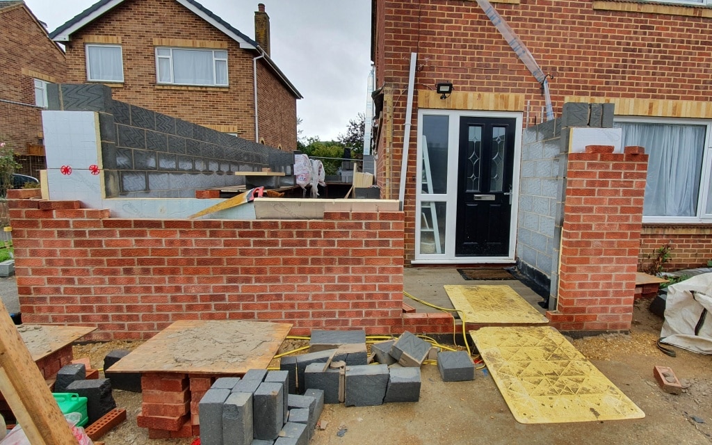 A partially built brick porch house extension in front of a house, with building materials, bricks, and yellow boards on the ground, and unfinished walls surrounding the front door.