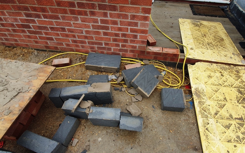 Several black concrete blocks, some broken, are scattered on the ground near a red brick wall. Yellow panels and cables are also visible at the building site for a house extension, where new bricks will soon join the existing structure.