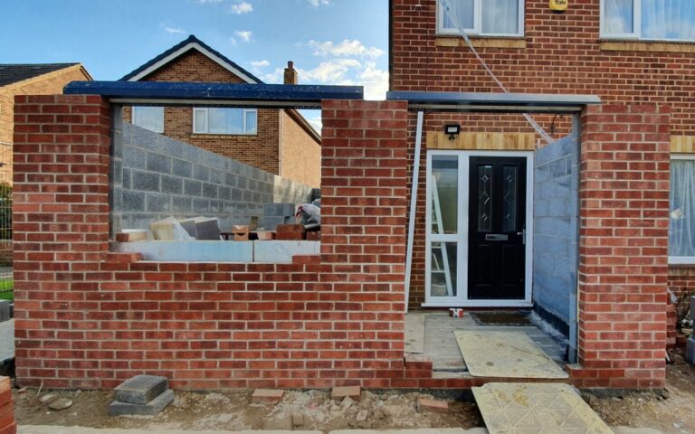 A house extension made of bricks is being built onto the front of a house. The structure features window and door openings, an unfinished roof, and building materials inside, with the main entrance door still accessible.