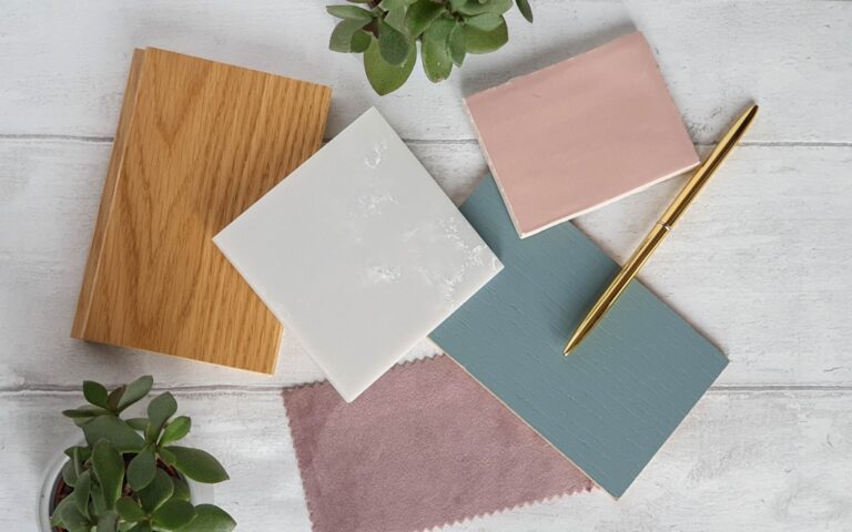 A flat lay of home decor samples for new kitchen inspiration: a wooden board, white and pink tiles, a blue painted sample, a gold pen, a pink fabric swatch, and green succulents on a white wooden surface.