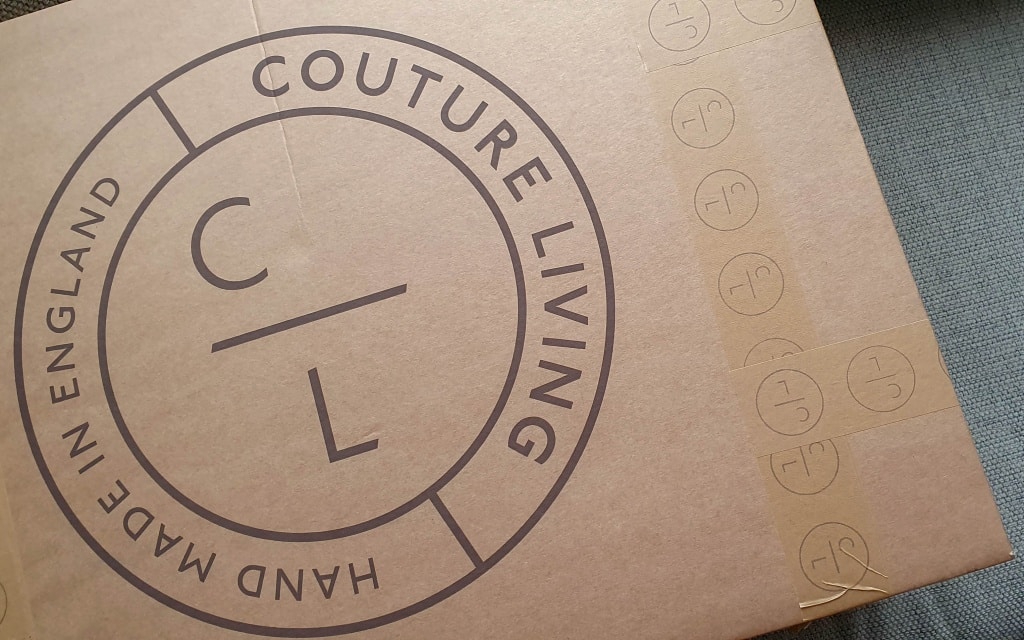 A brown cardboard box with the words Couture Living and Hand Made in England printed in a circular logo. Perfect for delivering made to measure curtains, the box is sealed with tape featuring repeating CL logos.