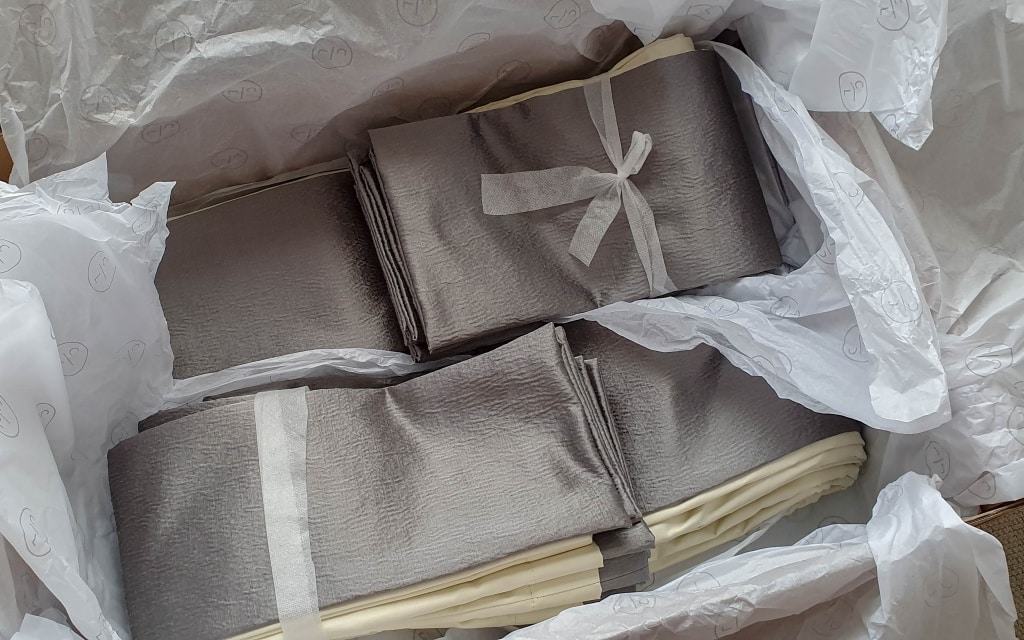Several neatly folded grey and cream-coloured fabric pieces, some tied with white ribbon, are arranged inside a box lined with crumpled white tissue paper—perfect samples for Couture Living made-to-order curtains for your bedroom.